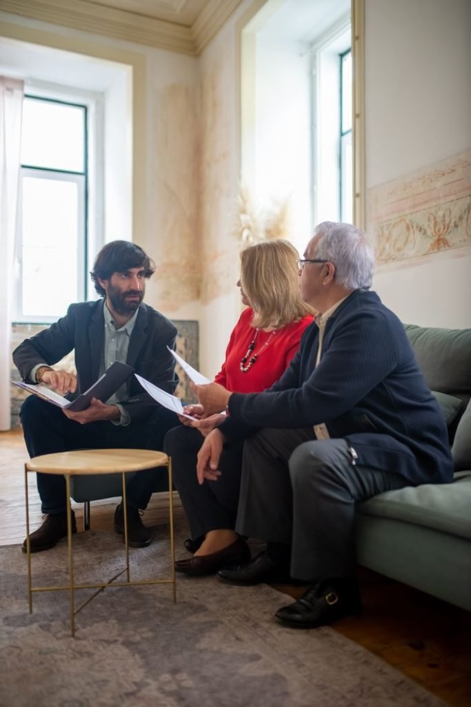 people-having-conversation-8439647 An advisor discussing financial documents with a senior couple indoors in a cozy living room setting.
